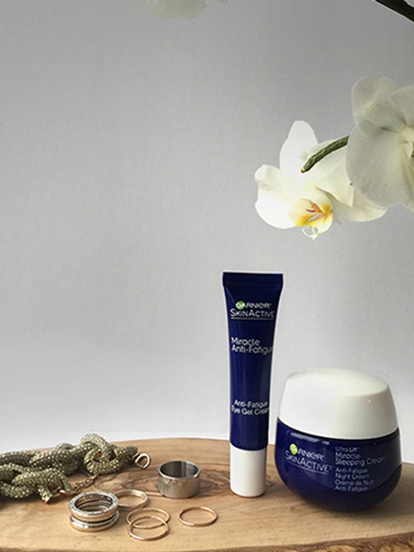 A tube of Miracle Anti-Fatigue Eye Gel Cream and a jar of Miracle Anti-Fatigue Night Cream sit on a wooden table beside various silver rings and a thick braided necklace on a wooden table beside a white orchid on a grey background.